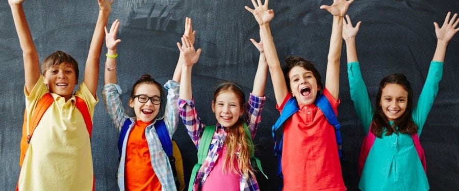 Three cheerful schoolchildren with backpacks raise their hands in excitement against a blackboard background, showcasing a joyful learning environment.