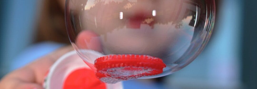 Close-up of a red candy or gummy on a clear surface, with a blurred background featuring a child's face. The image highlights the texture and vibrant color of the candy.