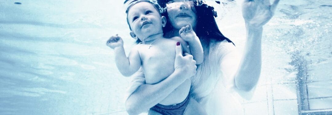 Mother and baby underwater, both smiling, capturing a joyful moment in a serene blue setting. The baby is held securely in the mother's arms, showcasing a loving bond.