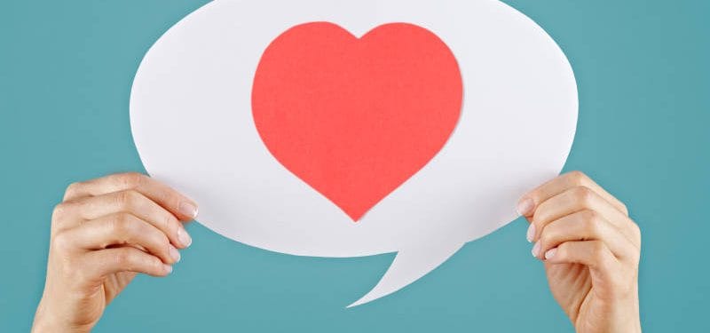 Hands holding a speech bubble with a red heart symbol, set against a teal background, representing love and communication.
