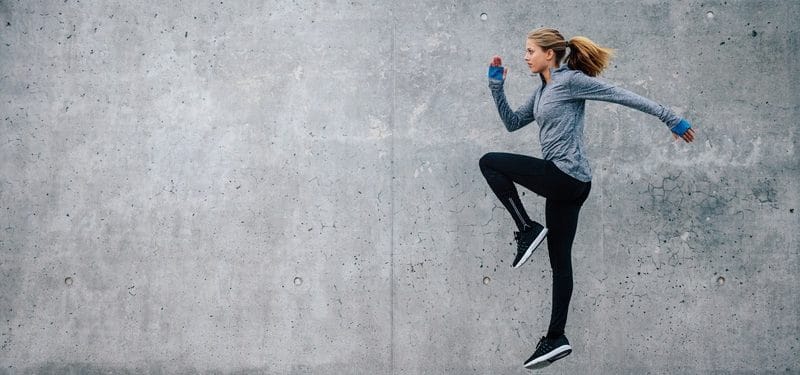 Athletic woman in activewear performing a running drill against a concrete wall, showcasing fitness and movement.