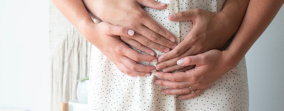 Close-up of multiple hands gently resting on a pregnant person's belly, symbolizing love and support during pregnancy. The hands are adorned with light nail polish, and the person is wearing a white dress with small polka dots.
