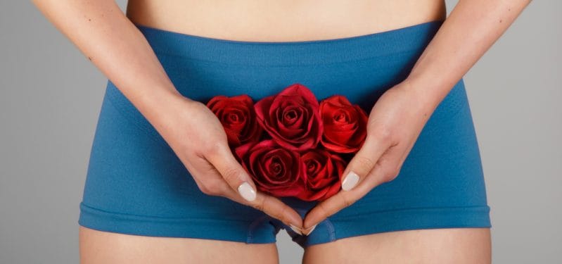 Person holding a bouquet of red roses in front of their lower abdomen, wearing blue shorts, symbolizing femininity and beauty.
