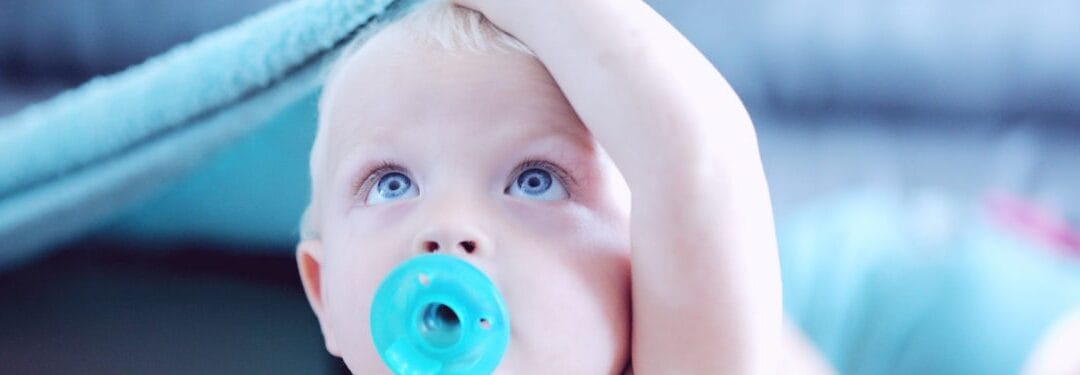 Baby with blue eyes holding their head, looking up while sucking on a turquoise pacifier.