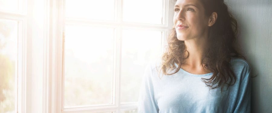 Young woman with curly hair standing by a sunlit window, looking thoughtfully outside, wearing a light blue top.