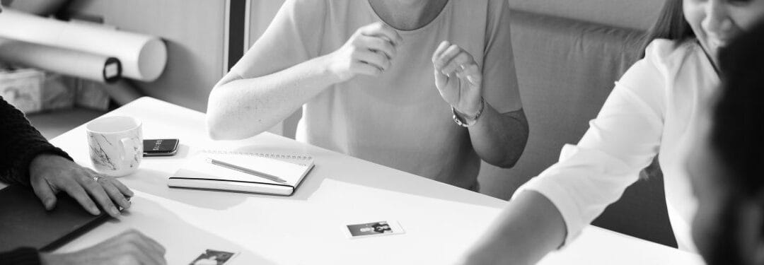 Person gesturing during a discussion at a table, with a notebook and a small photograph visible, in a black and white setting.