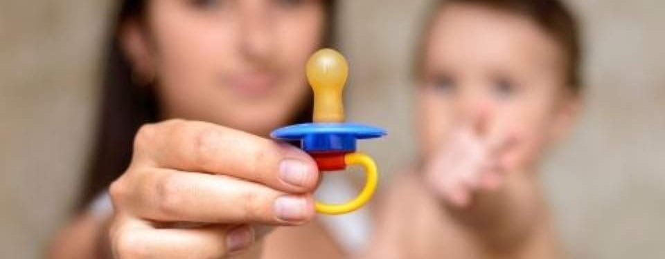 Close-up of a colorful baby pacifier being held by a caregiver, with a blurred baby in the background, illustrating essential baby care products.