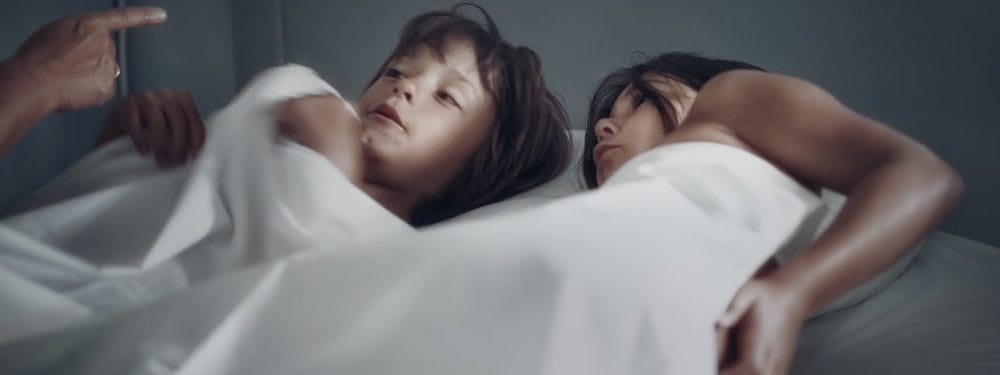 Two children lying in bed under white blankets, with one child speaking and the other listening attentively. The scene conveys a sense of intimacy and comfort in a cozy bedroom setting.