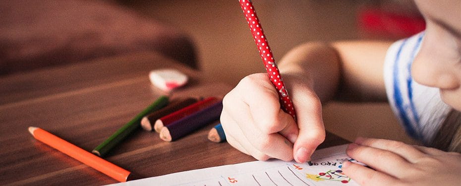 Child's hand holding a red polka-dotted pencil, drawing on a sheet of paper with colored pencils scattered on a wooden table.