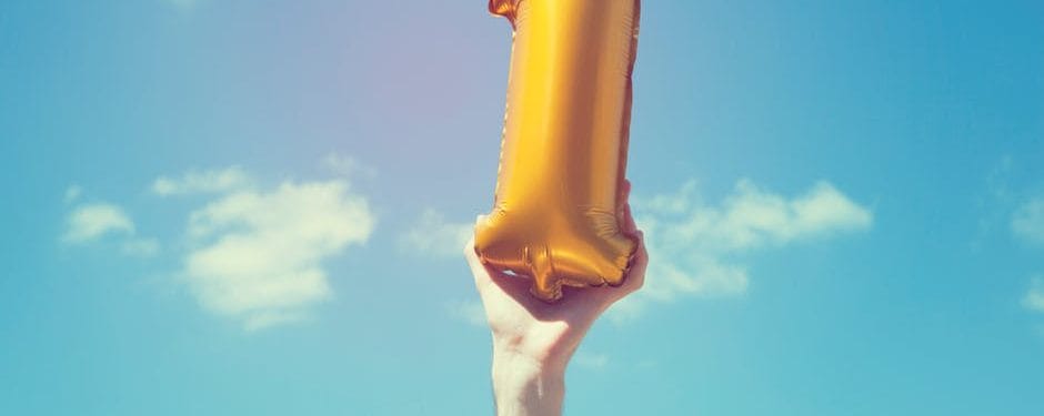 Hand holding a large gold number one balloon against a blue sky, symbolizing celebration and milestones.