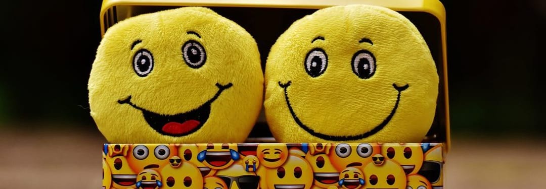 Two cheerful yellow emoji plush toys with smiling faces, displayed against a colorful background featuring various emoji patterns.