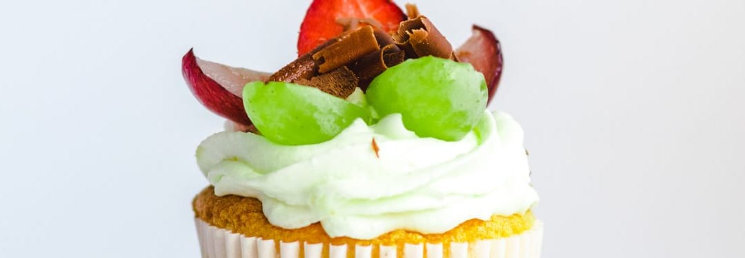 Colorful cupcake topped with whipped cream, chocolate shavings, green grapes, and fresh strawberries, showcasing a delightful dessert.