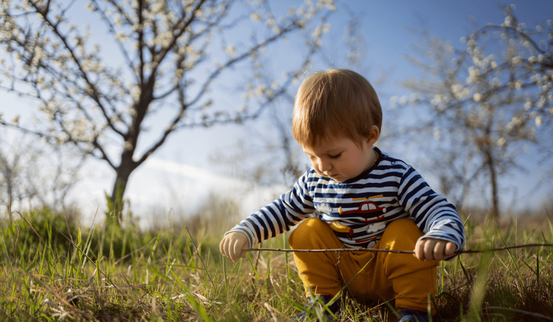 Young boy with short brown hair wearing a striped shirt and yellow pants, crouching in a grassy field, playing with a stick under a clear blue sky and flowering tree.