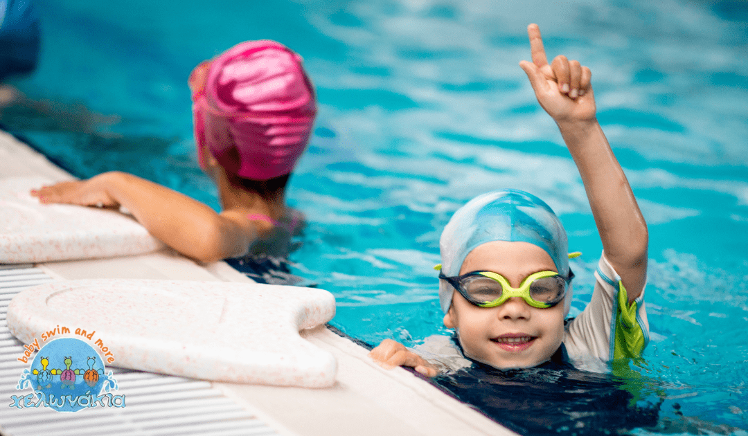 Two children enjoying swimming lessons in a pool, one wearing a light blue swim cap and goggles, smiling and raising a hand, while the other child in a pink swim cap swims nearby.
