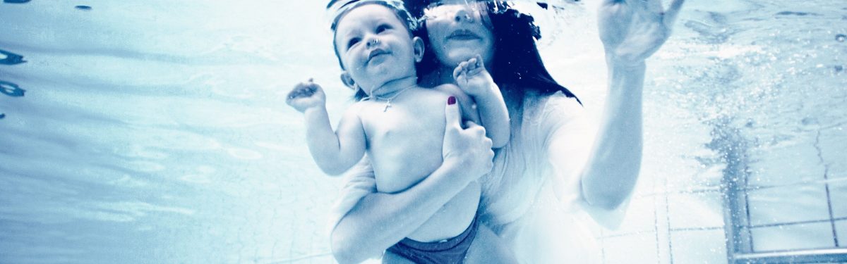 7-ofeli-tis-vrefikis-kolymvisis Mother and baby underwater, both smiling, capturing a joyful moment in a serene blue setting. The baby is held securely in the mother's arms, showcasing a loving bond.