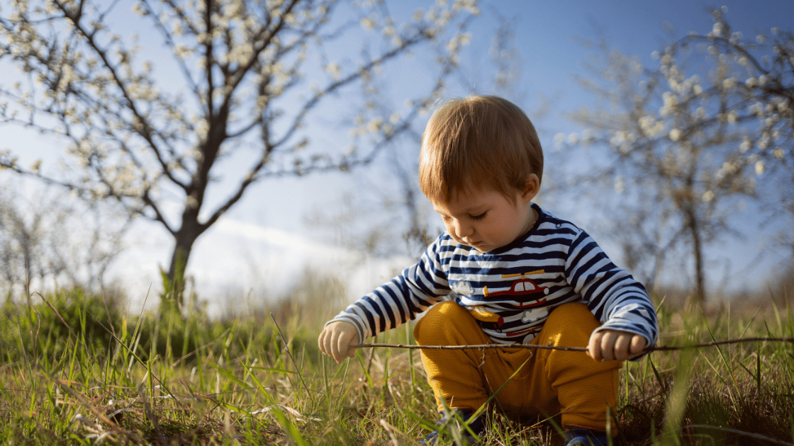drastiriotites-gia-paidia-2eton Young boy with short brown hair wearing a striped shirt and yellow pants, crouching in a grassy field, playing with a stick under a clear blue sky and flowering tree.