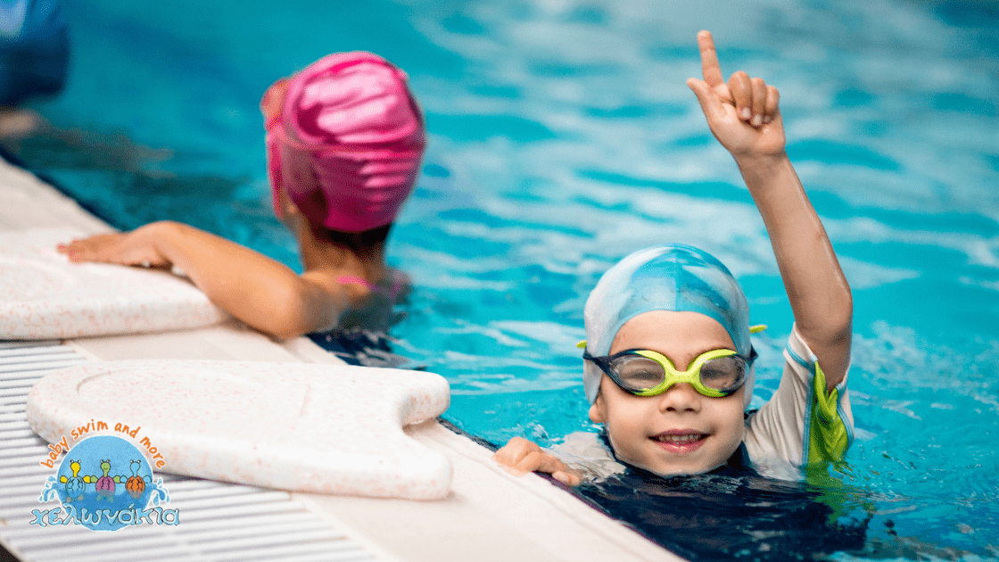 kolymvisi-gia-paidia-pote-na-ksekinisete Two children enjoying swimming lessons in a pool, one wearing a light blue swim cap and goggles, smiling and raising a hand, while the other child in a pink swim cap swims nearby.
