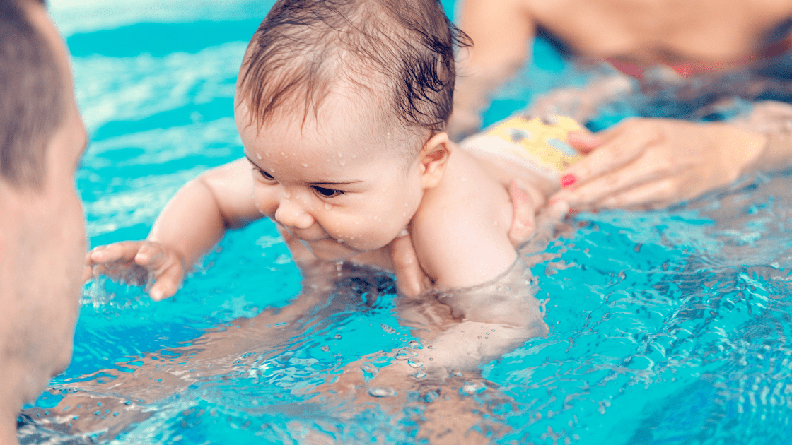 vrefiki-kolymvisi Baby swimming in a pool, exploring the water with playful curiosity while being supported by an adult.