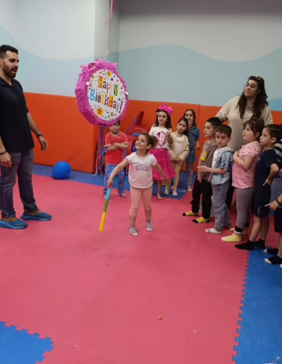 A young girl with a birthday-themed outfit prepares to hit a colorful pinata that says "Happy Birthday" at a children's party, surrounded by excited friends and family in a vibrant playroom.