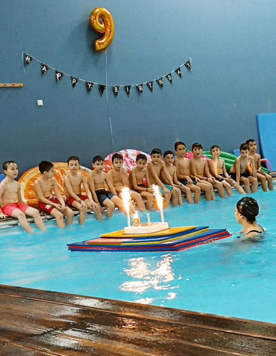 Children celebrating a birthday party at a pool, with a cake featuring candles on a colorful platform, surrounded by inflatable toys and festive decorations, including a large "9" balloon and a banner.