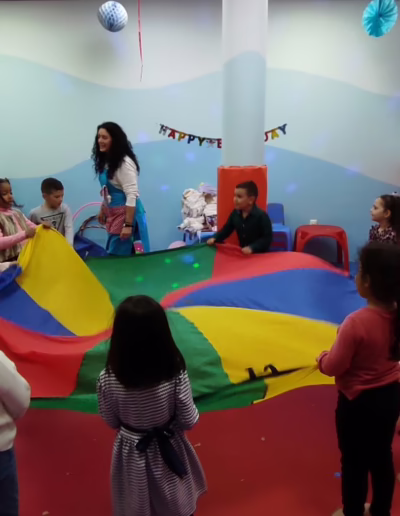 Children participating in a fun group activity with a colorful parachute in a playful indoor setting, guided by an instructor. The bright, cheerful environment is decorated for a celebration, with soft play areas and festive decorations.
