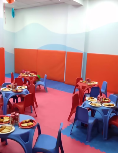 Colorful children's birthday party room featuring blue and red tables with plates of food, surrounded by playful balloons and padded walls for safety. A "Happy Birthday" banner decorates the wall, creating a festive atmosphere.