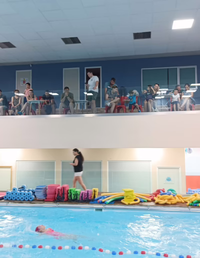 Indoor swimming pool scene with spectators observing from a balcony. Various colorful swimming aids are stacked by the poolside, while a swimmer in a pink swimsuit practices in the water. The environment is lively, showcasing a swimming lesson or event with engaged onlookers.